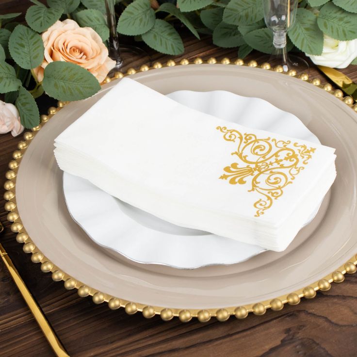 two white napkins sitting on top of a plate next to flowers and goldware