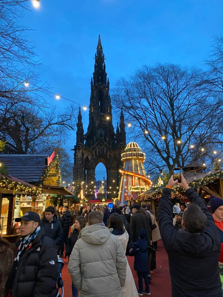 people are walking around an outdoor market with lights on the trees and buildings in the background