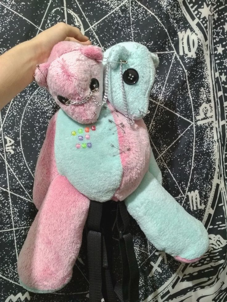 a pink and blue teddy bear sitting on top of a black bag next to a person's hand