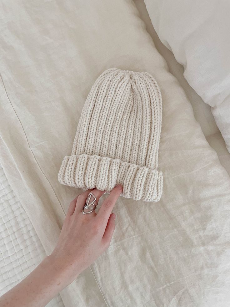 a hand is holding a knitted hat on top of a white bed with pillows