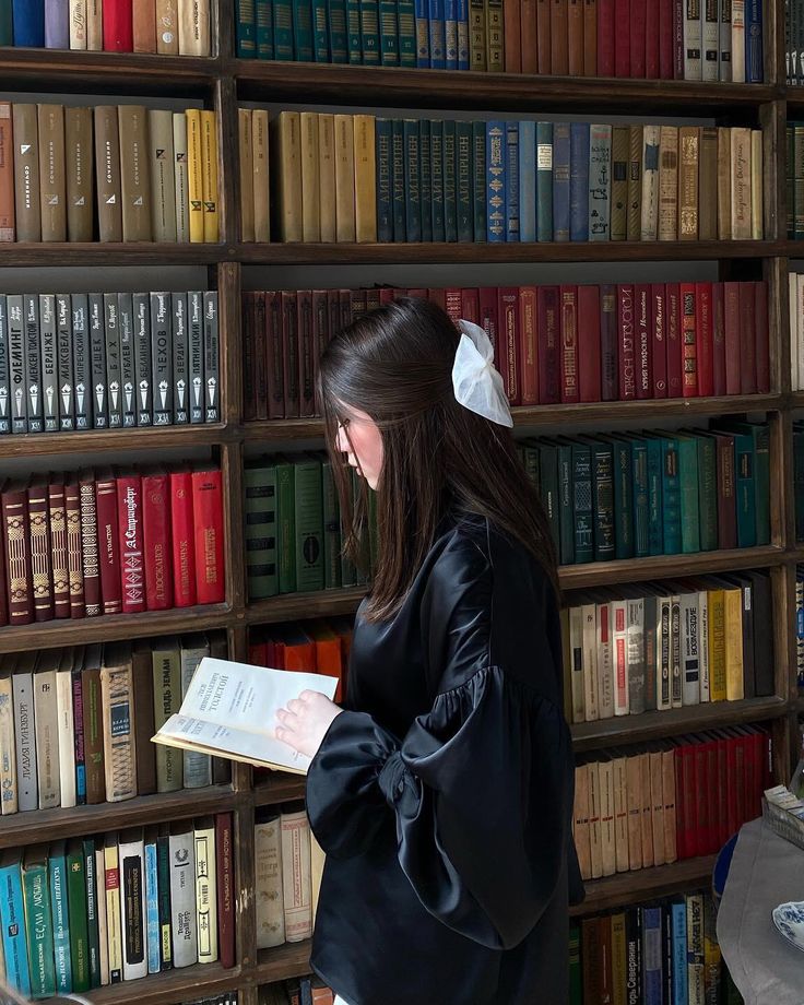 a woman standing in front of a bookshelf holding a book and looking at it