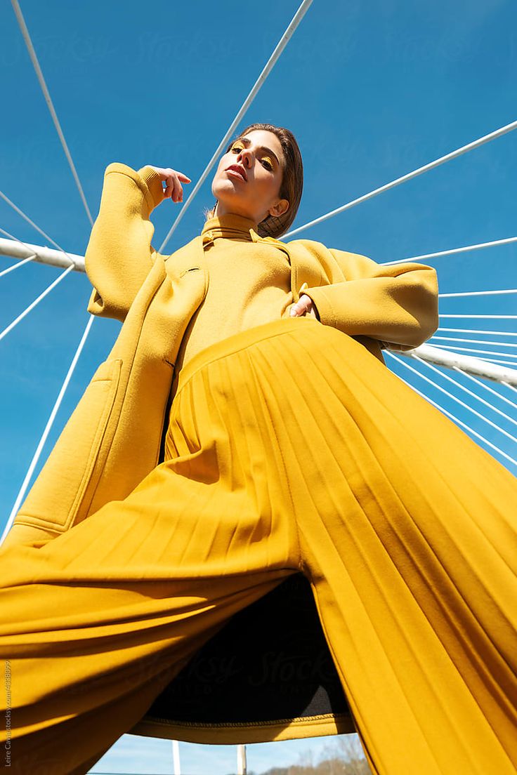 a woman in a yellow dress leaning on a white pole with blue sky behind her