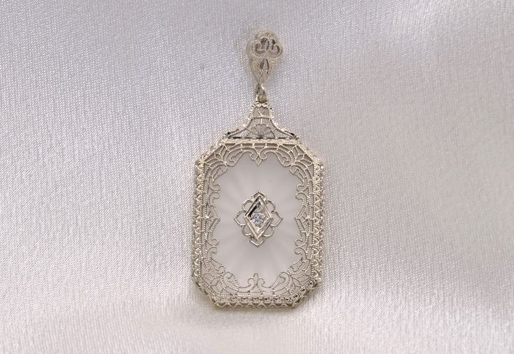 The 21st Street: Antique Art Deco Camphor Glass Pendant with Diamond. The pendant centers on a filigree section set with old cut round natural diamond. The frosted sun burst camphor glass is rectangular in shape and is framed by elaborate and intricate filigree finished with fine milgrain. A filigree top and bail complete the look of this 14 karat white gold Art Deco-era camphor glass pendant. The pendant is photographed on a chain for display purposes only. Inquire about purchasing a chain to a Luxury Rectangular Pendant Jewelry For Wedding, Luxury Rectangular Pendant Wedding Jewelry, Elegant White Rectangular Jewelry, White Gold Rectangular Pendant For Wedding, Elegant Rectangular Pendant Jewelry For Wedding, Antique Diamond Intaglio Jewelry, Elegant White Rectangular Pendant Jewelry, Classic Wedding Jewelry With Rectangular Pendant, Elegant Rectangular Jewelry With Single Cut Diamonds