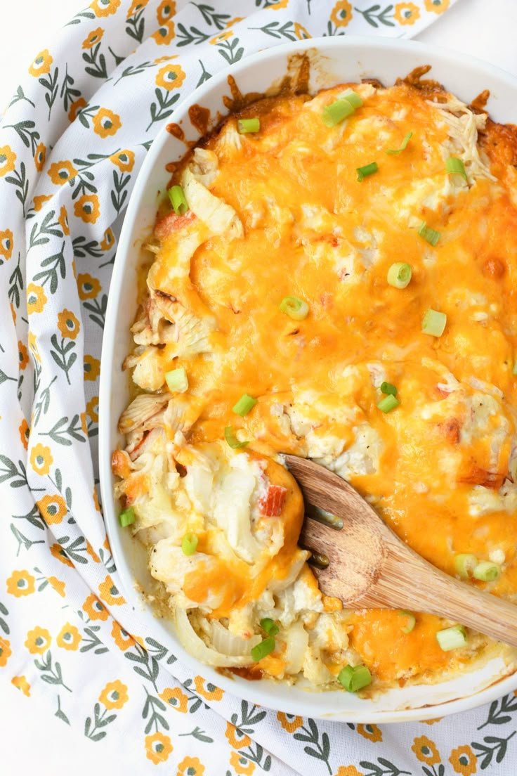 a casserole dish with chicken and vegetables in it on a floral cloth next to a wooden spoon