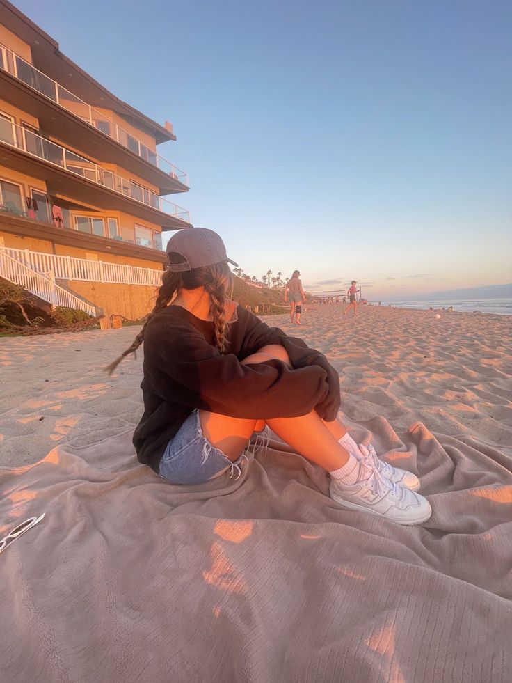 a woman sitting on top of a sandy beach next to an apartment building at sunset