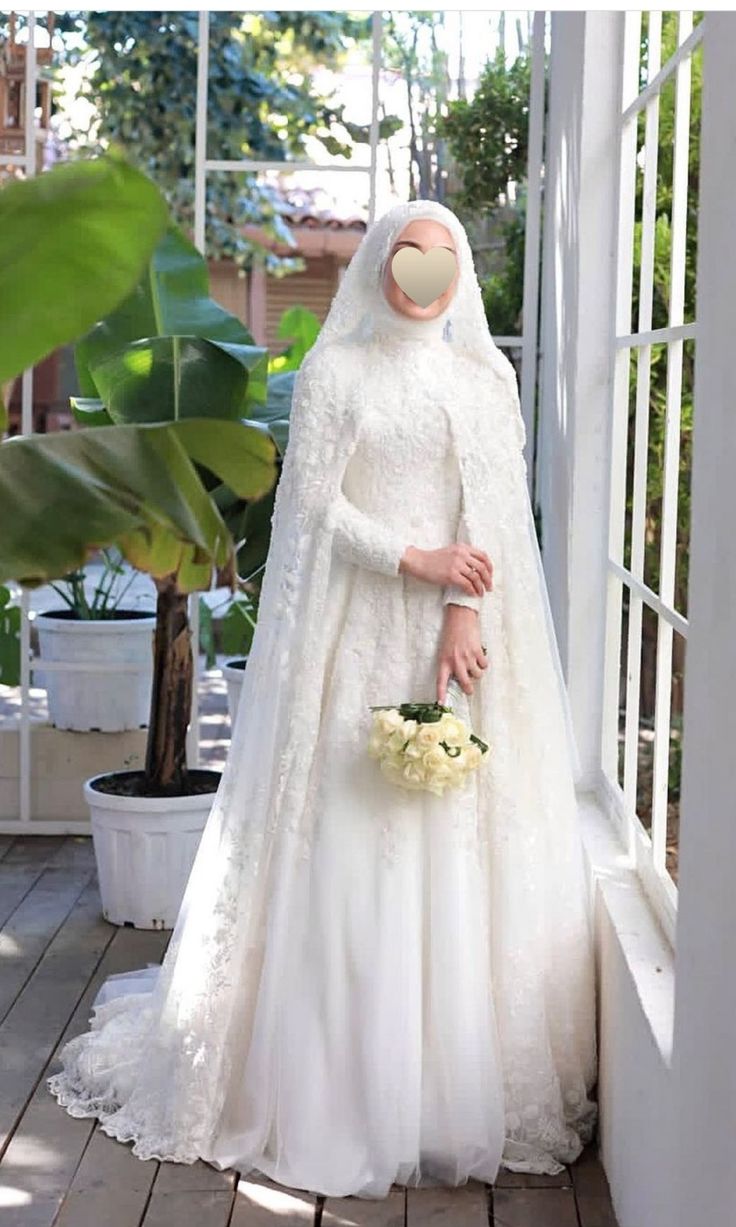 a woman in a white wedding gown and veil holding a flower bouquet standing on a porch