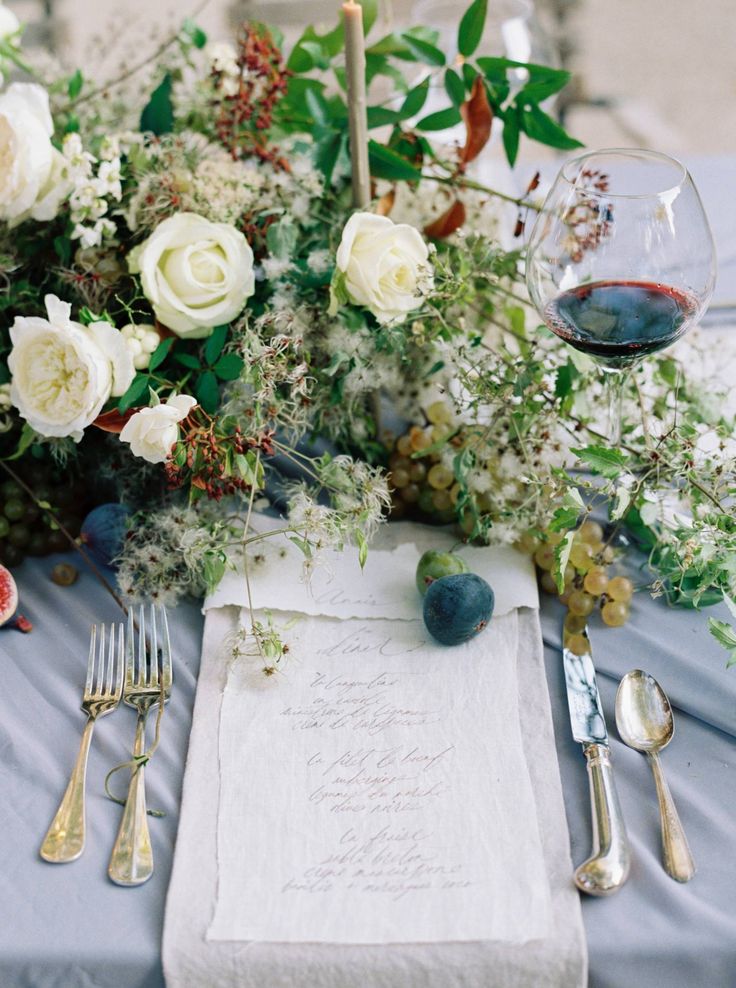 a table topped with white flowers and silverware next to a menu on top of a blue cloth