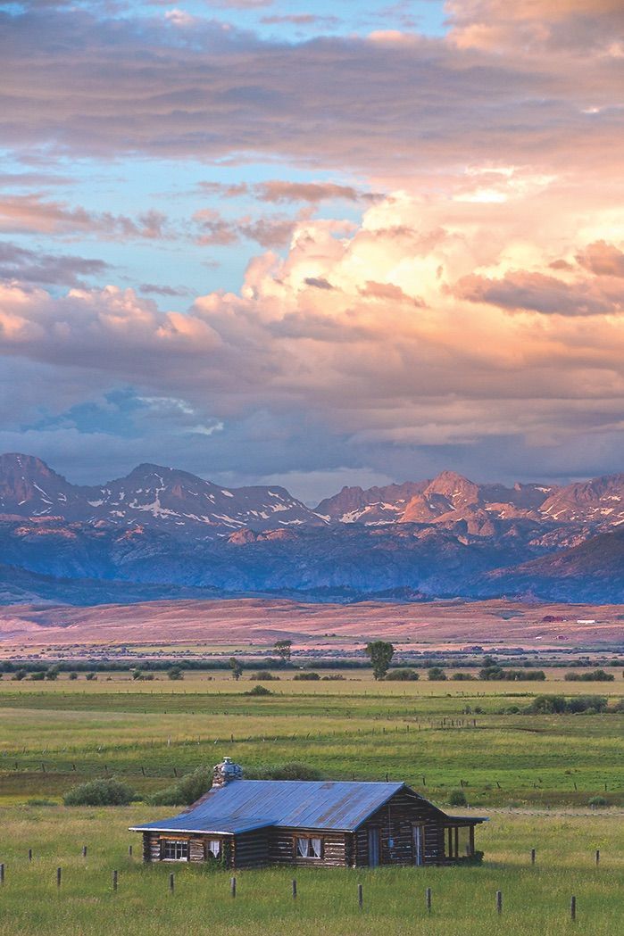 an old barn sits in the middle of a field with mountains in the back ground