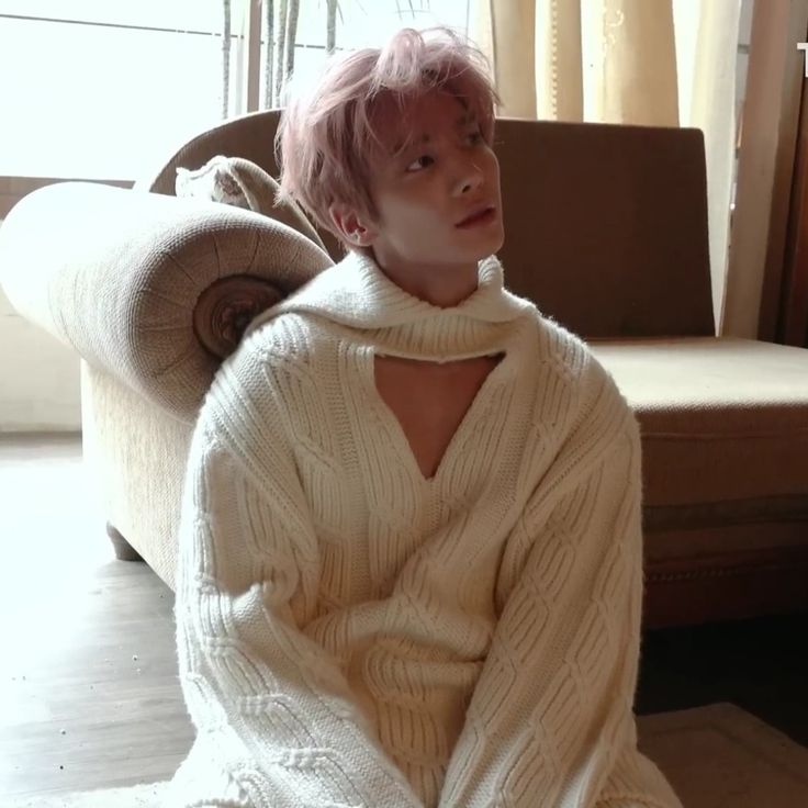 a person with pink hair sitting on a couch wearing a white sweater and black pants