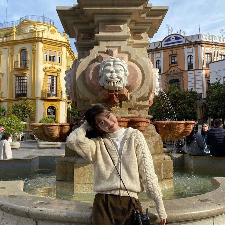 a woman standing in front of a fountain with a handbag on her shoulder and wearing a white sweater