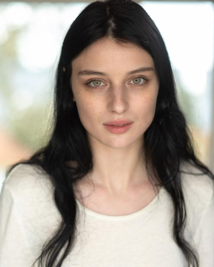 a woman with long black hair and blue eyes looks at the camera while standing in front of a window