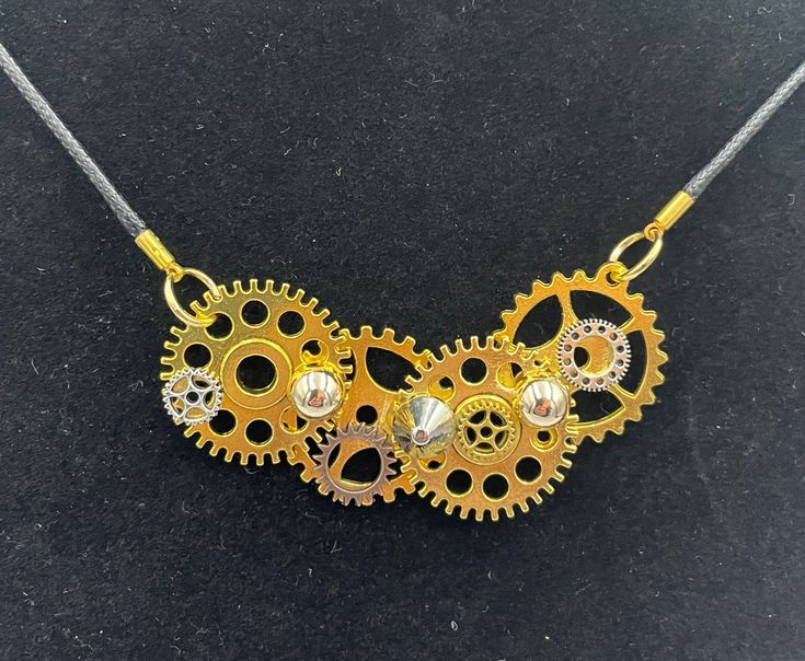 Unique handmade steampunk style necklace, designed and created by engineers in Scotland. A really special gift for someone who appreciates original design, or as a quirky treat for yourself.  The combination of the linked gears on the black cord results in a dainty minimalist necklace with a striking industrial feel.  The main part of the necklace is made from gears, cemented together and then combined using unique looking screw back rivets. The rivets are domed and spiked, leading to an asymmetrical look. The gear combination is approximately 70mm long and 35 wide from the furthest away points. It is attached to a waxed black cord. The full length is 52cm and the cord closure is a gold tone lobster claw, ensuring the closure blends sleekly into the design. The necklace is delivered in an Steampunk Gold Jewelry With Adjustable Chain, Adjustable Gold Steampunk Necklaces, Adjustable Gold Steampunk Necklace, Gold Steampunk Jewelry With Adjustable Chain, Steampunk Metal Necklace With Adjustable Chain, Steampunk Style Metal Necklace With Adjustable Chain, Steampunk Adjustable Necklace With Soldered Details, Gold Steampunk Metal Necklaces, Steampunk Necklace With Adjustable Metal Chain