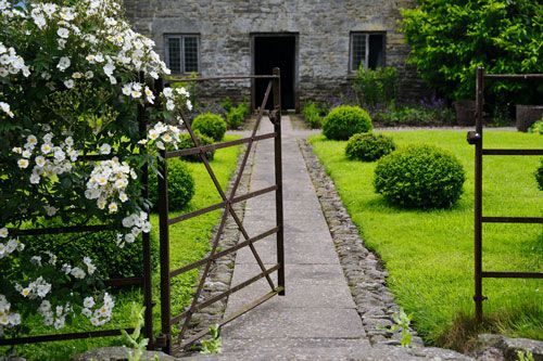 an open gate leading to a stone building with white flowers in the foreground and green grass around it