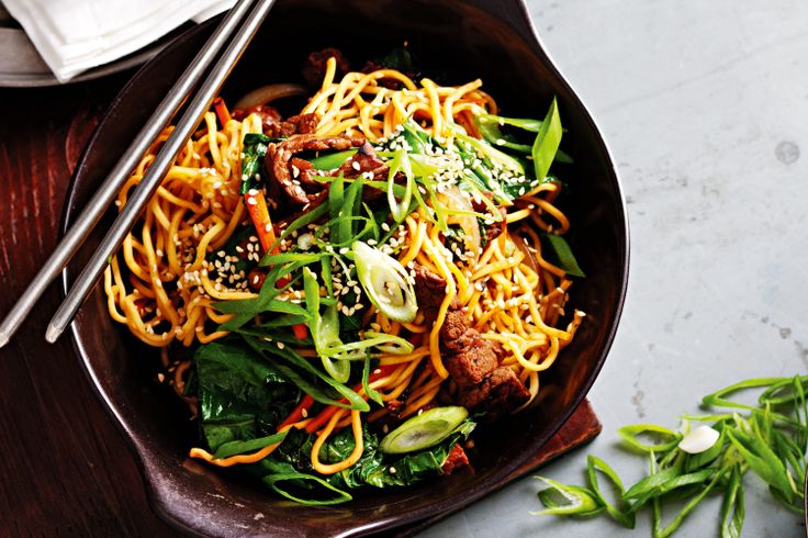 a bowl filled with noodles and vegetables on top of a wooden table next to chopsticks