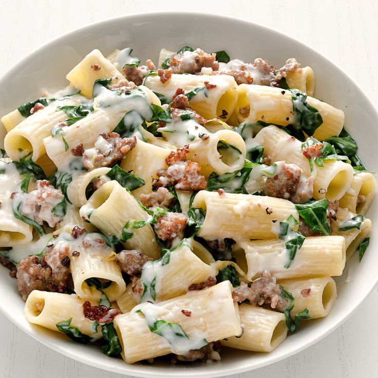 a white bowl filled with pasta, meat and spinach covered in cream sauce on top of a wooden table