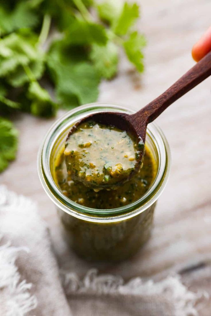 a wooden spoon is in a jar filled with green sauce and cilantro leaves