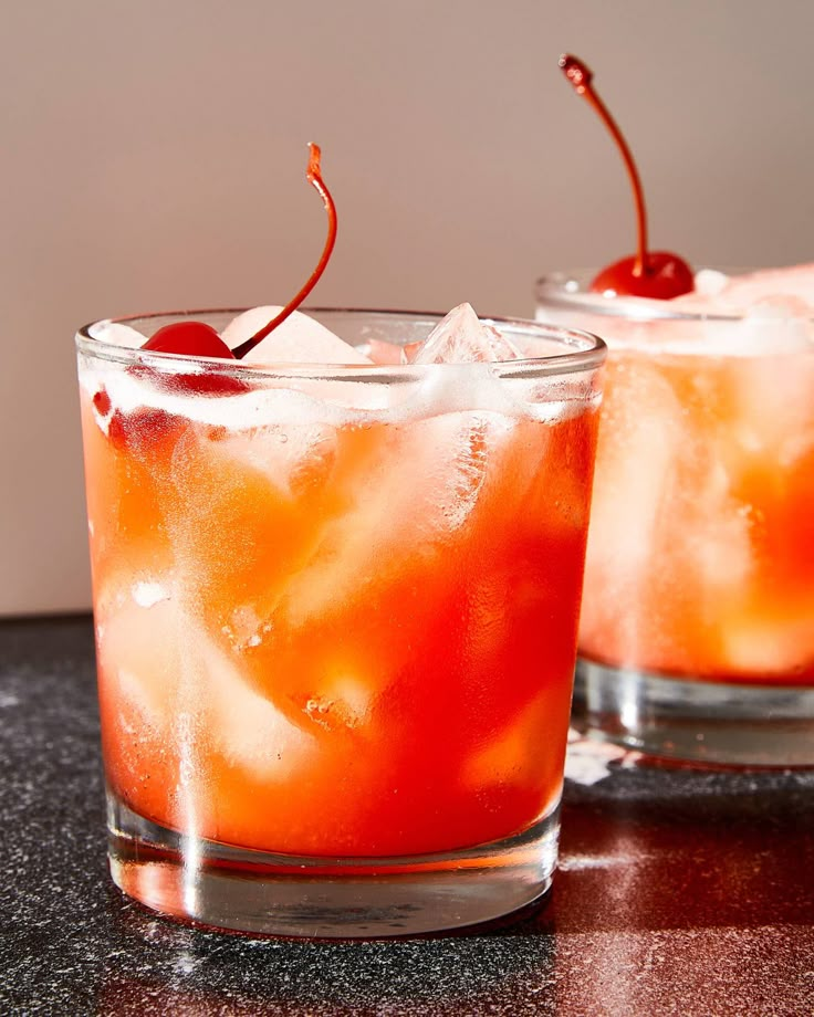 two glasses filled with ice and cherries on top of a black counter next to each other