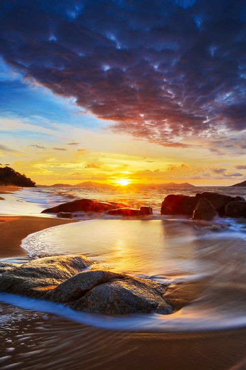 the sun is setting over the ocean with rocks in the foreground and waves on the shore