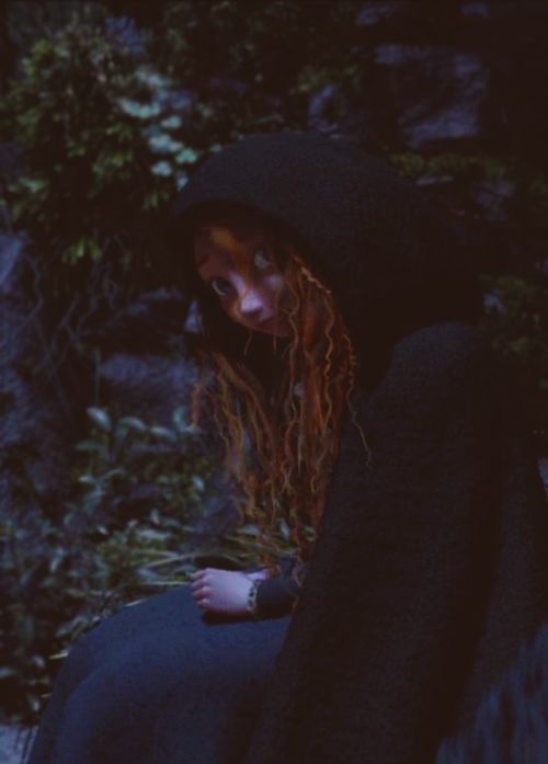 a woman with long red hair wearing a black cloak and sitting on a rock in the woods