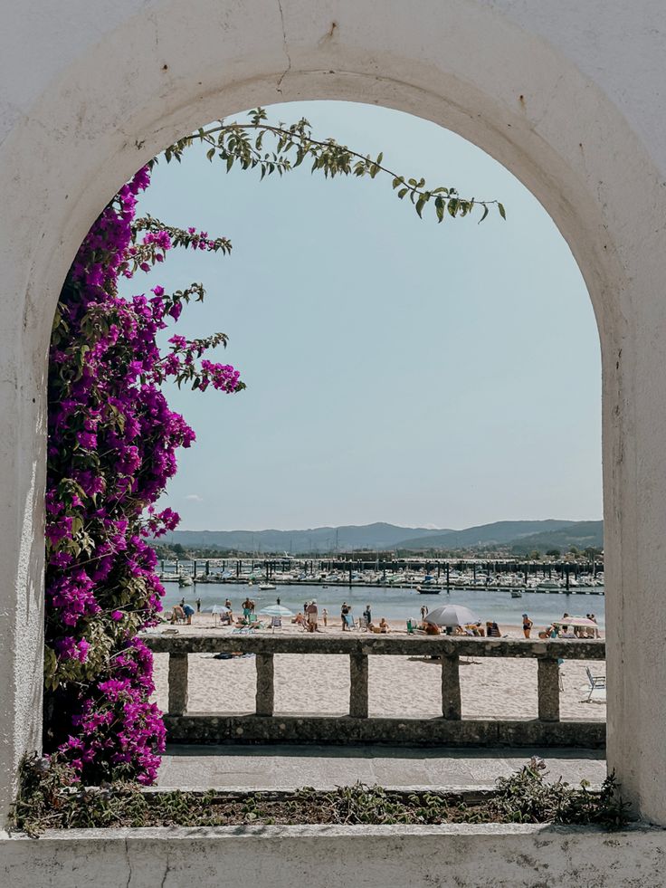 an archway with purple flowers on the beach and people in the water behind it,