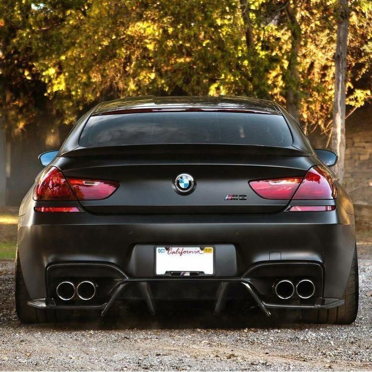 the back end of a black bmw car parked in front of some trees and bushes