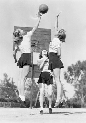 three young women playing basketball in an old black and white photo, one jumping up to the basket