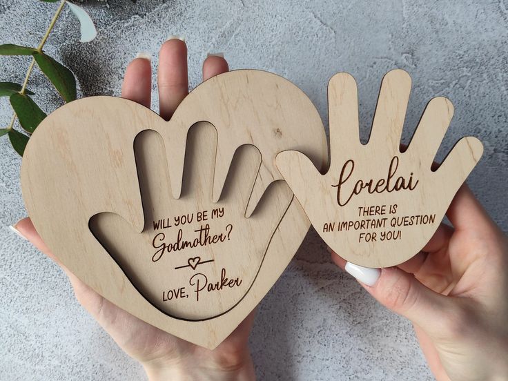 two wooden heart shaped handprints that say, will you be my godmoter?