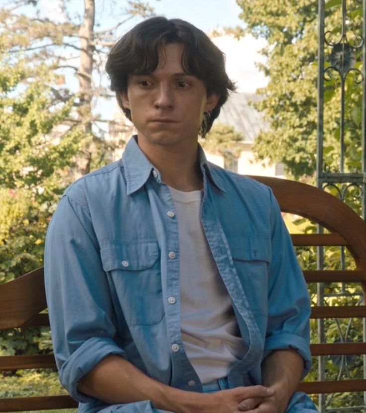 a man sitting on top of a wooden bench in front of some trees and bushes