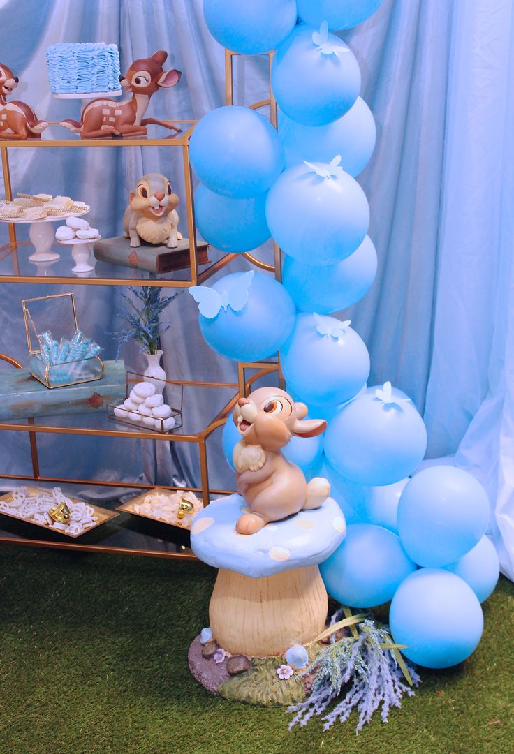 a table with blue balloons and stuffed animals on the shelves, including a cake stand