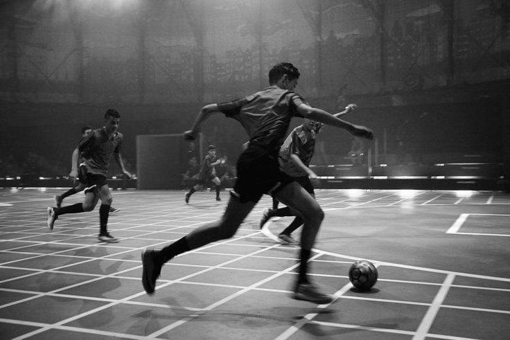 three men are playing soccer in an indoor arena with lights shining on the floor and one man is about to kick the ball