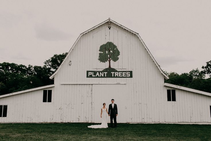 a bride and groom standing in front of a large white barn with a plant tree sign on it