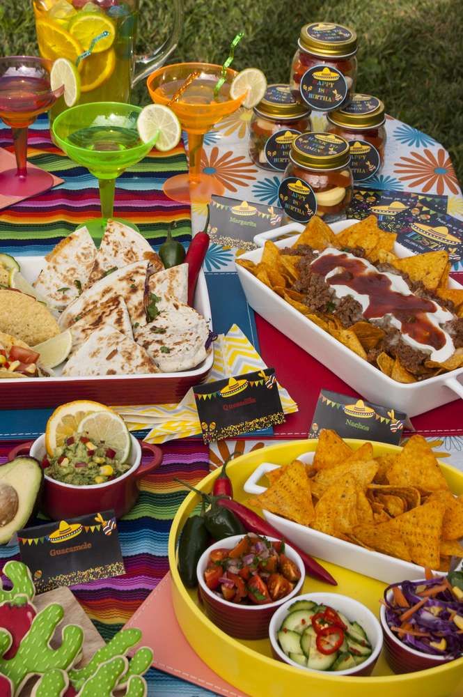 an assortment of mexican food on a colorful table cloth with drinks and snacks around it