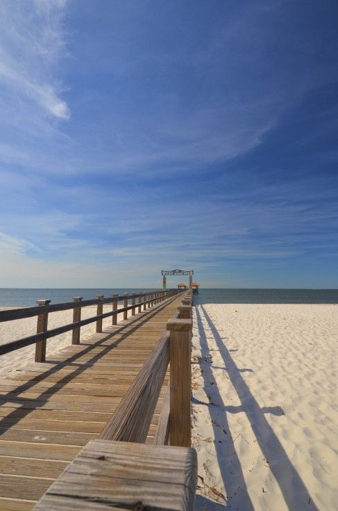 a long wooden boardwalk stretches out into the distance on a beach with white sand and blue sky