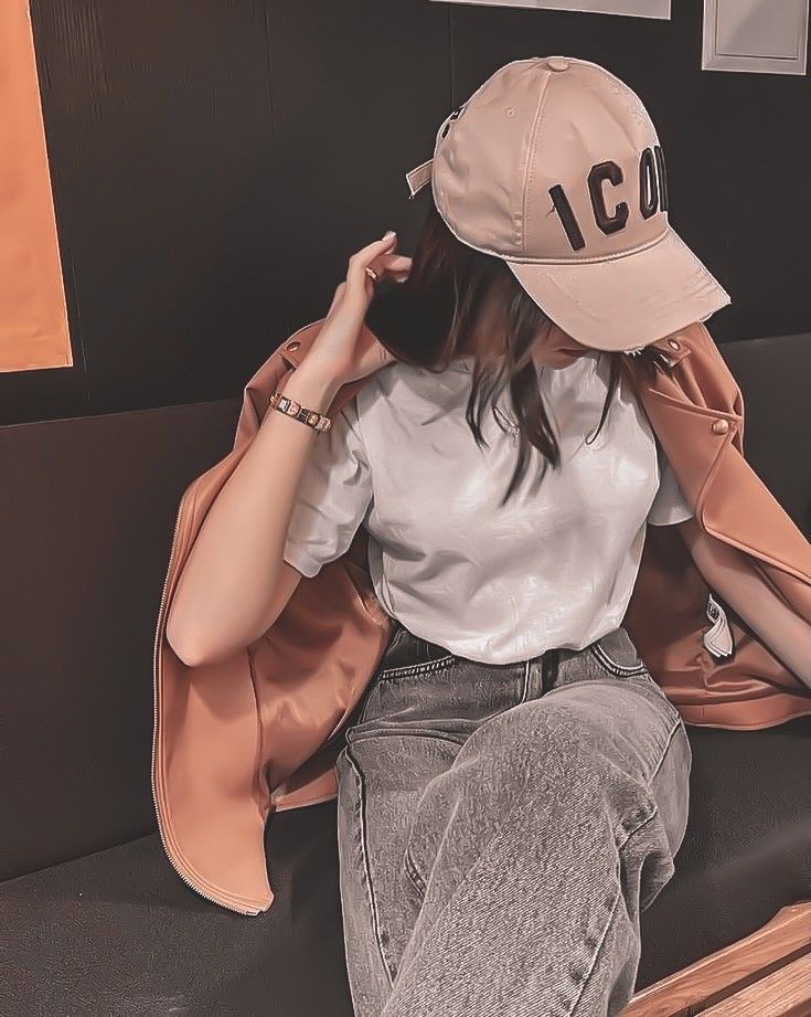 a woman sitting on top of a wooden bench wearing a baseball cap and grey jeans