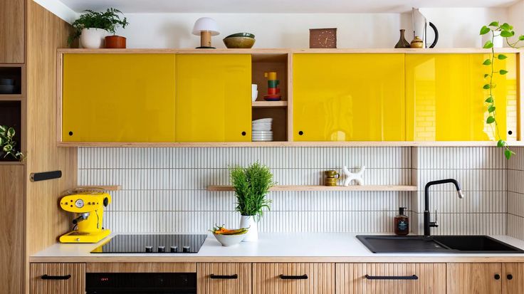 a kitchen with yellow cupboards and white counter tops, plants on the shelf above the sink