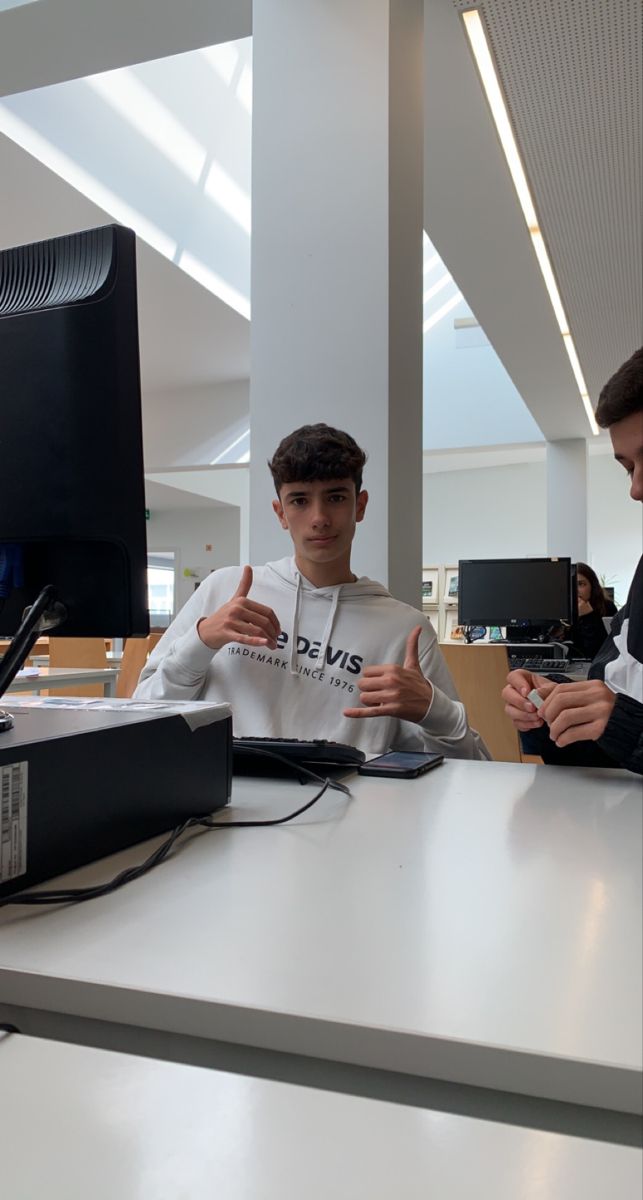 two young men sitting at a table with computers on the desk in front of them