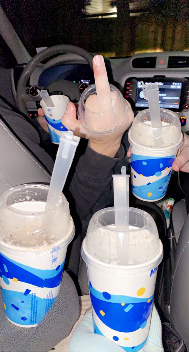 three plastic cups with drinks in them sitting on the dashboard of a car and someone holding up their peace sign