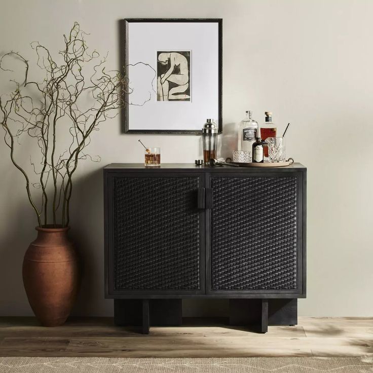 a black cabinet sitting next to a vase with some plants in it and a framed photograph on the wall