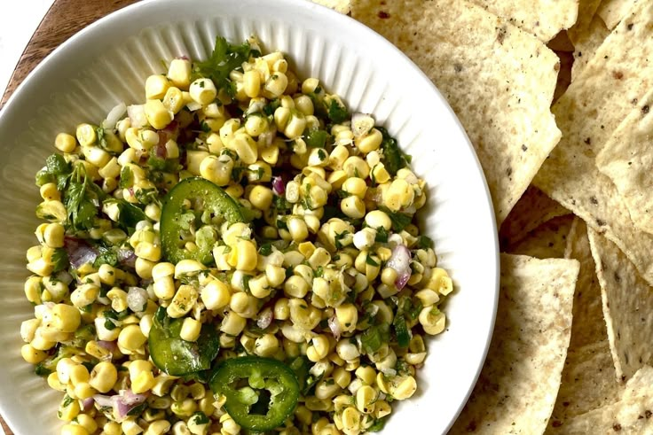 a white bowl filled with corn and vegetables next to tortilla chips on top of a wooden table