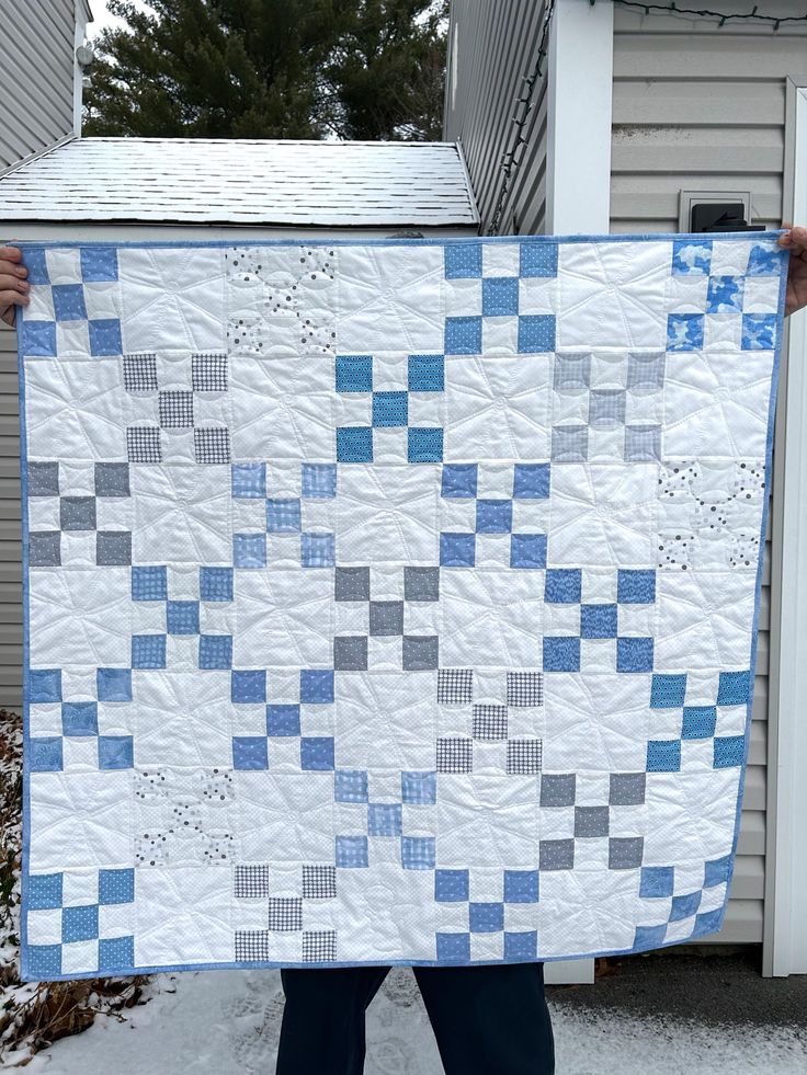 a person holding up a blue and white quilt in front of a house with snow on the ground