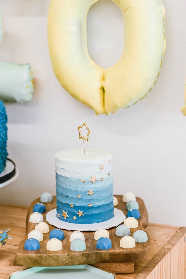 a blue and white cake sitting on top of a wooden cutting board next to balloons