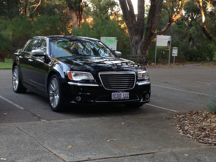 a black car parked in a parking lot next to some trees and bushes on the side of the road