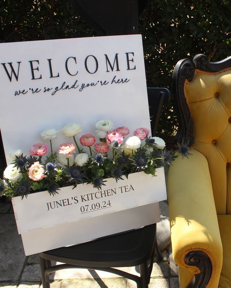 a welcome sign sitting on top of a yellow chair next to a flower pot filled with white and pink flowers