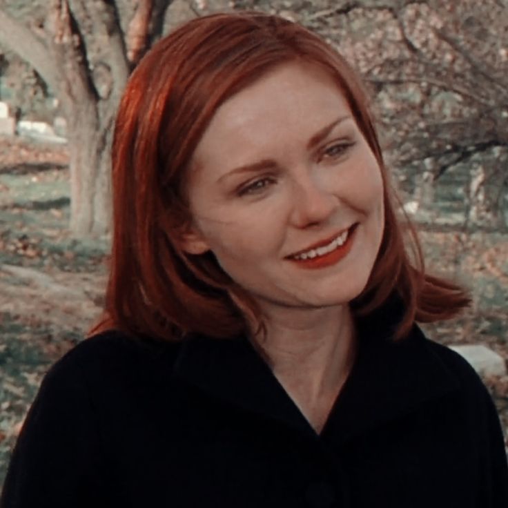 a woman with red hair smiling at the camera in front of some trees and rocks