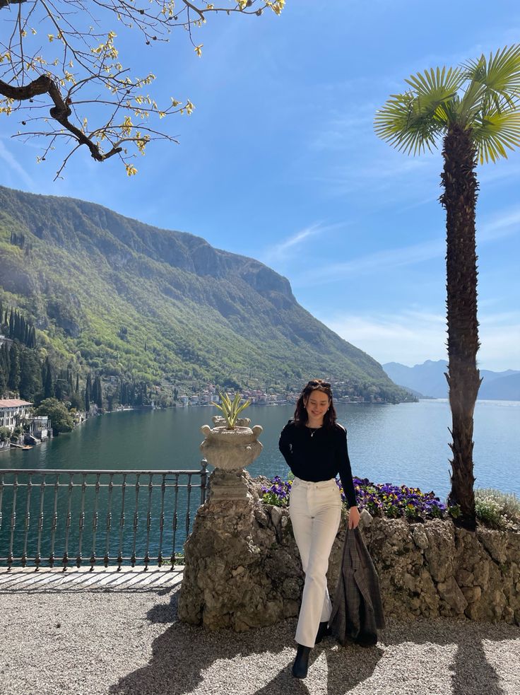 a woman standing next to a palm tree on top of a stone wall near the ocean