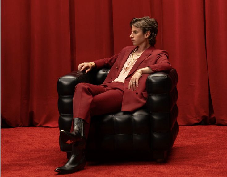 a man in a red suit is sitting on a black chair with his legs crossed