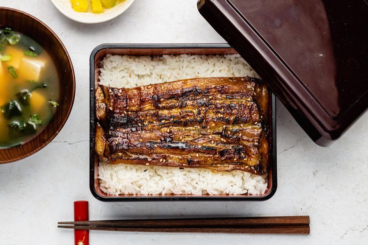 an assortment of food including rice, meat and chopsticks on a white table