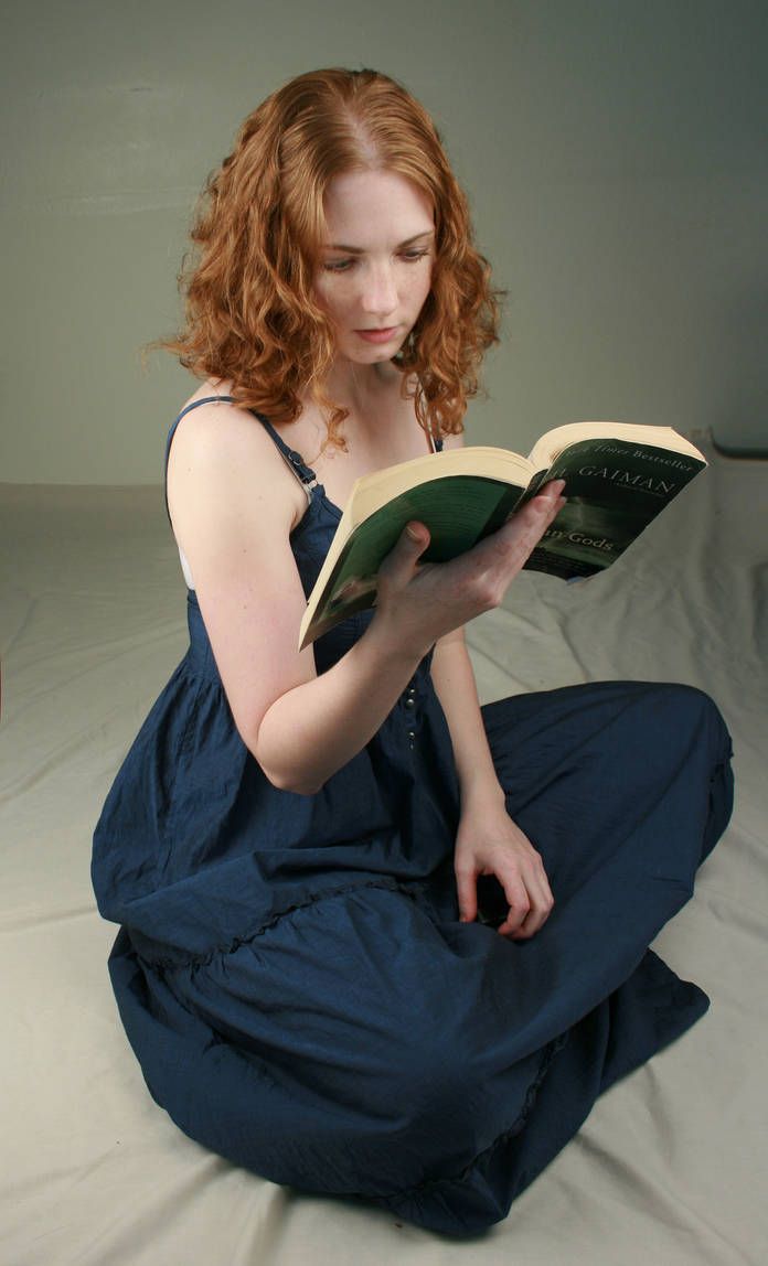 a woman sitting on a bed reading a book with red hair and blue pants,