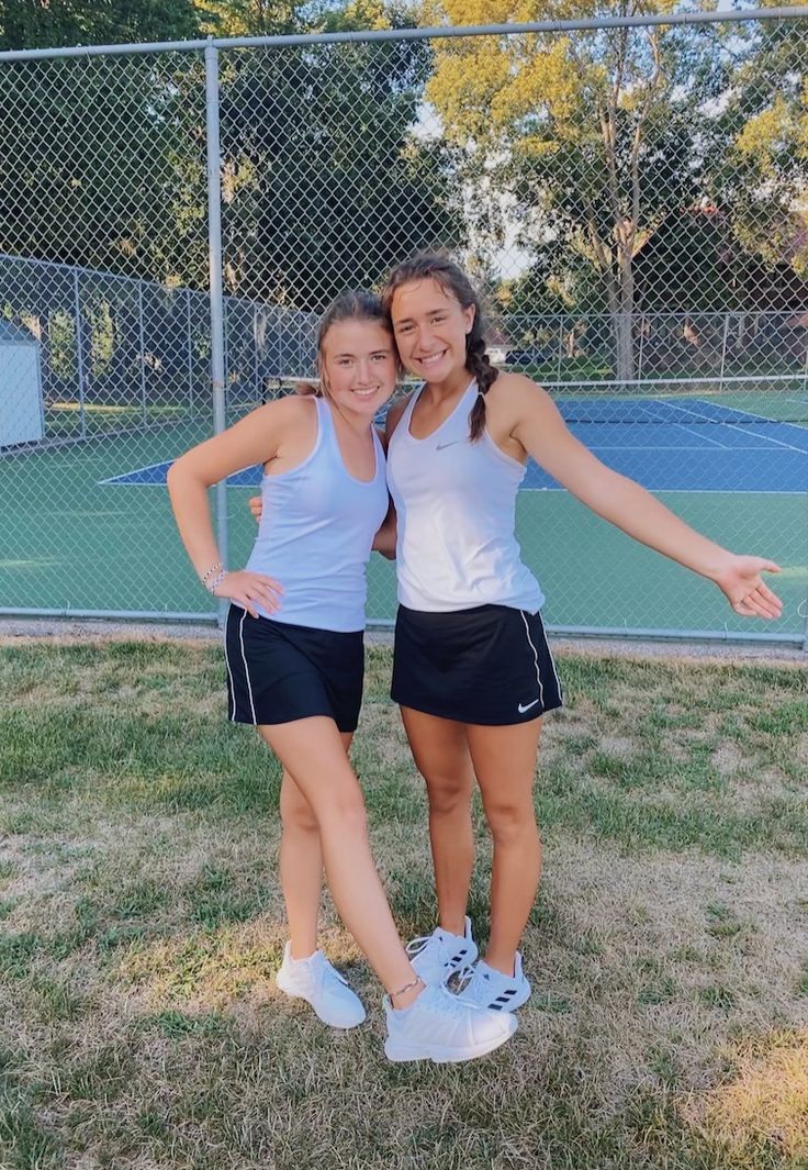 two young women standing next to each other in front of a tennis court