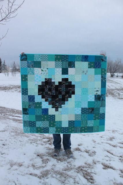 a person standing in the snow holding up a blue and black heart shaped patchwork quilt